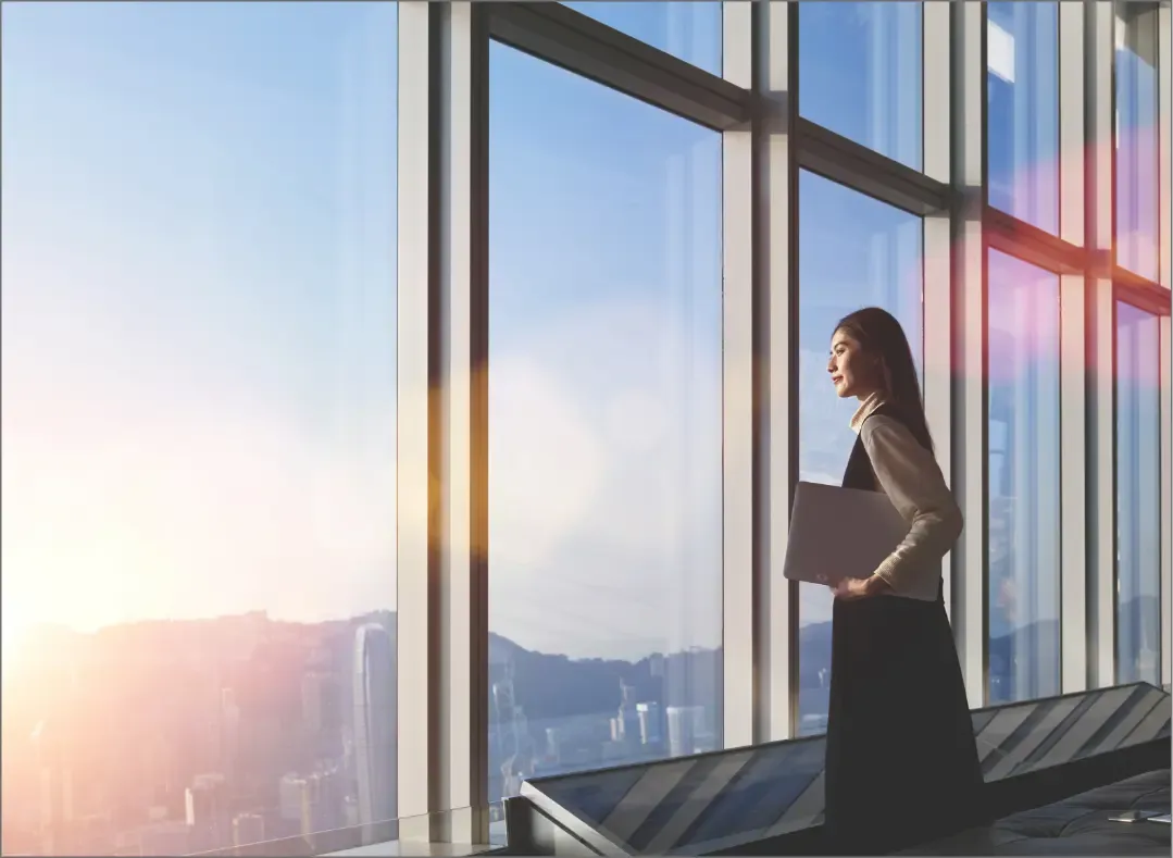A professional woman standing by large office windows holding a laptop