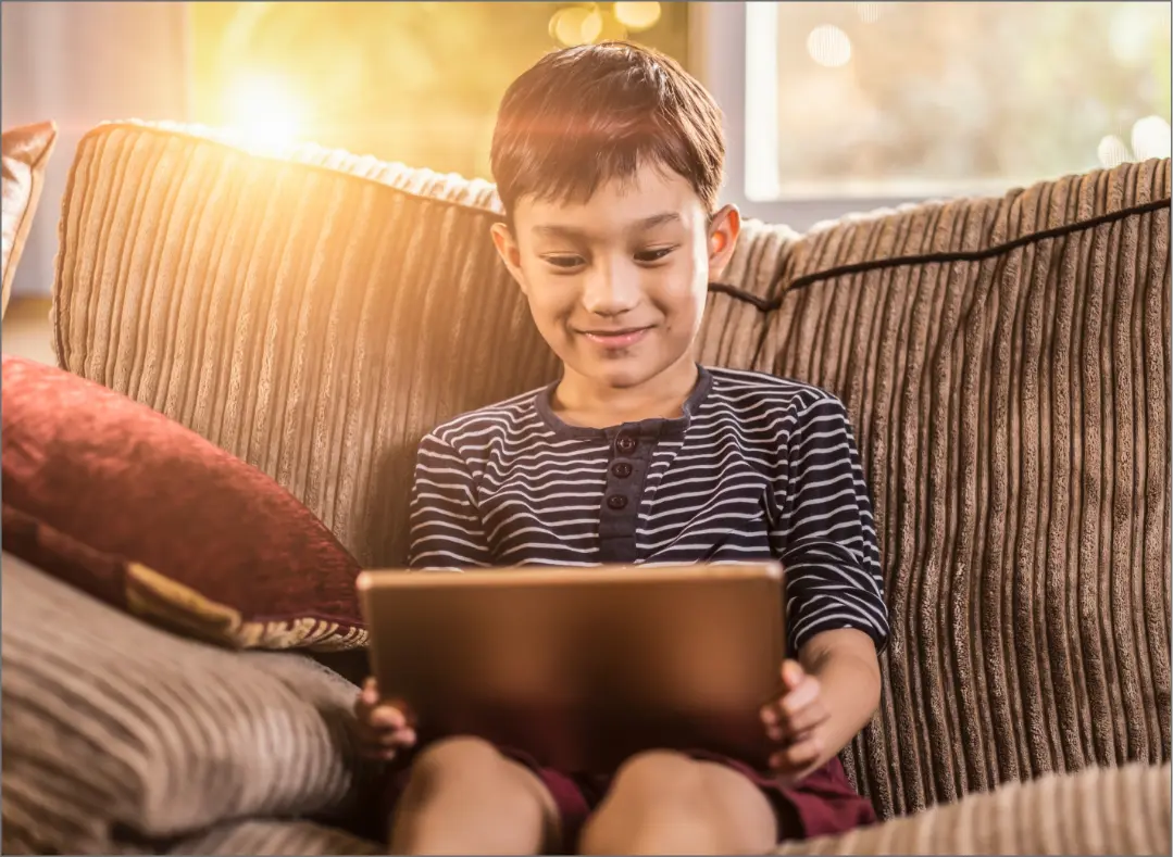 A smiling child using a tablet on a couch, showcasing digital entertainment and educational solutions by Trinergy.