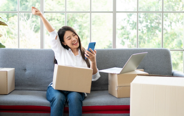 A happy woman celebrates a successful online shopping experience while sitting on a couch with boxes and a laptop, using her smartphone.